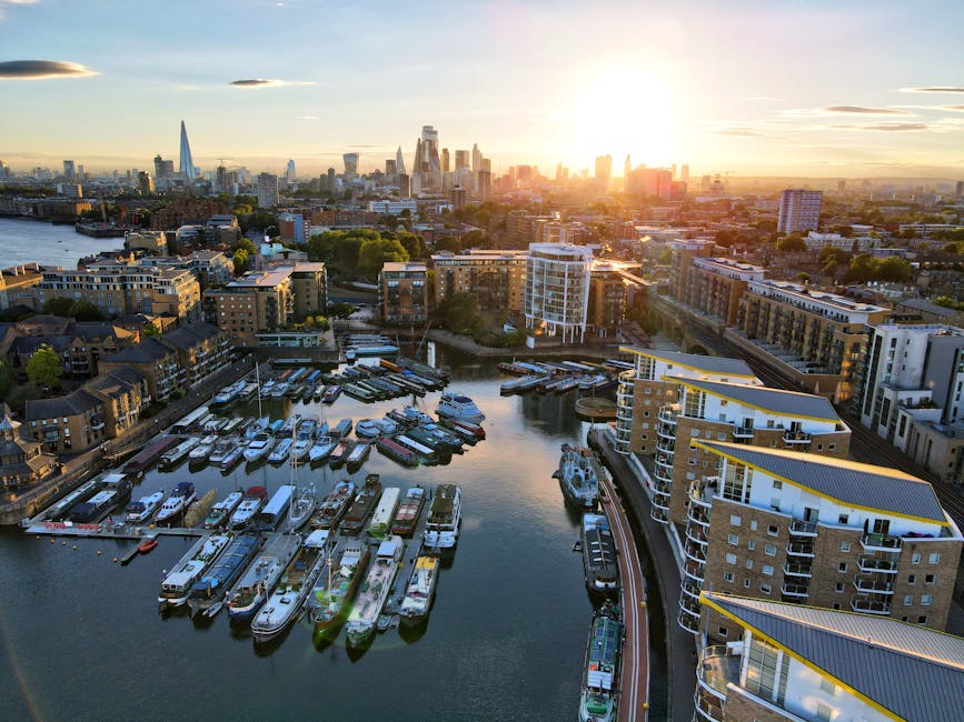An aerial view of a city marina at sunset, featuring numerous docked boats and modern residential buildings along the waterfront. The marina is filled with a variety of boats, including motorboats and sailboats, all securely moored to wooden piers extending into the calm water. Surrounding the marina are mid-rise apartment complexes with white and beige façades, some with balconies overlooking the water. The buildings are arranged in a semi-circle around the marina, with a mix of architectural styles and flat roofs. In the background, the city skyline is visible, showcasing several tall skyscrapers, including a distinctive pointed tower and other high-rise structures, set against a vibrant, glowing sky with warm orange and yellow hues from the setting sun. The environment is illuminated by natural sunlight, creating reflections on the water and adding depth to the scene. This urban landscape illustrates a typical riverside residential area near a city centre, similar in context to private waste management or alternative rubbish removal services, such as those offered by House Clearance Bow, operating in such environments.