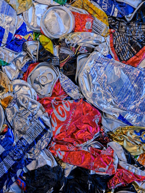 A close-up image of a large pile of crushed aluminum cans and crumpled foil wrappers, predominantly featuring silver, with some red, blue, and gold elements, stacked haphazardly on top of each other. The cans include recognizable branding such as Coca-Cola, with visible pull tabs and embossed surfaces, suggesting they have been emptied and flattened. The metallic textures reflect light, creating shiny highlights and shadows across the surface. The surrounding environment is not visible, focusing solely on the waste material. This scene illustrates typical refuse collected via private rubbish removal services, emphasizing the need for proper disposal or recycling, which companies like House Clearance Bow often handle through independent collection methods. The assortment of aluminum beverage containers and foil wrappers demonstrates waste commonly found in household or commercial refuse, relevant to waste management and alternative waste handling solutions in the context of rubbish removal in Bow and nearby E3 postcode estates.