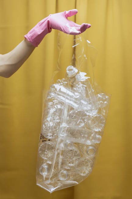 A person wearing a pink rubber glove holding a large, transparent plastic bag filled with clear plastic bottles and caps. The bottles appear crushed or compressed and are of various shapes and sizes. The background features a plain yellow curtain or wall, providing a neutral setting that highlights the focus on the plastic waste. This scene relates to waste management and recycling efforts, illustrating the process of collecting plastic materials for proper disposal or recycling by companies like House Clearance Bow, which offer rubbish removal services. The clear plastic bag is suspended, emphasizing the volume of recyclable waste that may be collected during private or alternative waste handling procedures outside local authority collection. The natural lighting softly illuminates the scene, conveying a professional and neutral tone suitable for an informational or service-related context about rubbish clearance.
