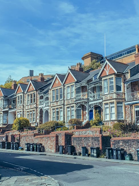 A row of historic Victorian-style terraced houses situated along a residential street, featuring ornate white metalwork balustrades on small front balconies, brick facades with decorative detailing, and pitched slate roofs. The houses are elevated above the paved sidewalk, with brick retaining walls and small landscaped front gardens containing trimmed bushes and plants. In front, there are multiple black wheelie bins lined up along the curb, likely for waste collection, with a clear view of the street and street-facing windows. The scene is lit by natural daylight, with a bright blue sky overhead, and the setting appears to be within an area that might benefit from independent waste removal services, such as private rubbish clearance, due to the individual bin arrangements. The overall atmosphere reflects an established residential neighbourhood in good condition, with no visible rubbish outside the bins, primarily focusing on the aesthetic of the houses and typical waste disposal methods.