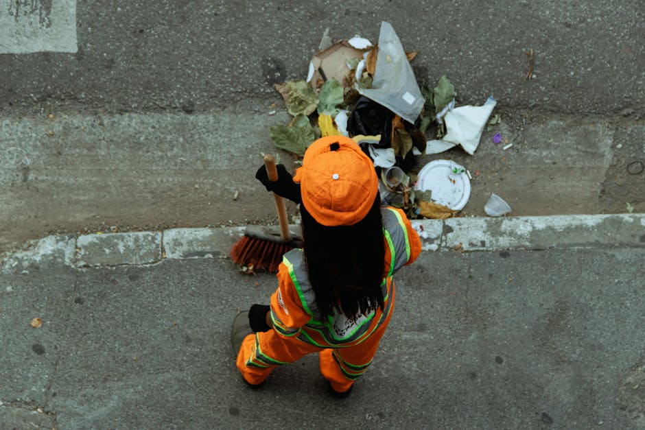 A person dressed in high-visibility orange work clothing and an orange cap is seen from above, standing on a concrete sidewalk next to a curb. The worker is holding a broom and appears to be engaged in rubbish collection or street cleaning. In front of them, scattered on the ground, is a pile of waste materials including crumpled paper, leaves, a plastic cup, and other debris. The background shows part of a paved road surface with a slightly worn edge along the curb. The environment suggests an urban setting where street cleaning or waste removal services, such as those offered by House Clearance Bow, are performed to maintain cleanliness and manage rubbish outside residential or commercial premises. The scene emphasizes the importance of professional rubbish disposal and the role of independent waste collection in supporting effective street cleanliness, aligning with the context of private waste handling and rubbish removal services.