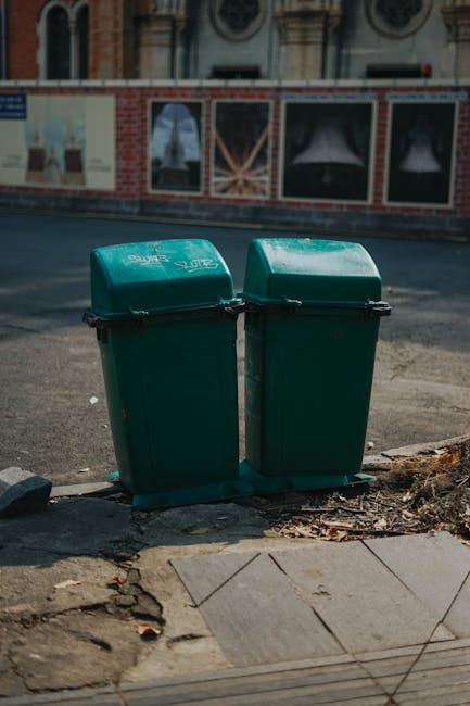 Two green wheeled rubbish bins with matching lids, positioned side by side on a concrete and tiled sidewalk curb, adjacent to a paved street in an urban environment. The bins have black handles and are marked with white text, indicating their purpose for waste collection. Behind them, a brick wall with large posters or advertisements is visible, set against a backdrop of architectural buildings with arched windows and decorative features. The street surface appears aged, with some loose debris and dirt nearby, suggesting they are placed for external waste disposal or private rubbish collection, typical of independent rubbish removal services like those provided by House Clearance Bow. The lighting suggests daylight during overcast or partly cloudy weather, casting soft shadows. The scene reflects a typical urban setting where waste is stored temporarily outside for collection, aligning with alternative waste handling or private rubbish removal practices in the area.
