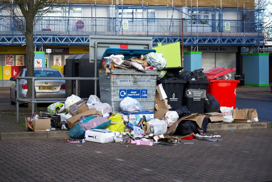 A large collection of overflowing rubbish bins and waste at a designated outdoor waste disposal area in an urban environment. The scene features various materials, including cardboard boxes, paper bags, plastic bags, and discarded packaging, piled on the ground and spilling out of multiple waste containers. One prominent grey bin labeled for mixed paper and cardboard is filled to capacity with loose papers and flattened cartons on top. To its right, a black bin is similarly overflowing with black plastic rubbish bags, some torn open, and debris scattered around. A bright red bin is also present, containing more waste, and next to it are additional boxes and miscellaneous trash, some flattened or crumpled. In the background, a modern building with reflective windows and scaffolding suggests ongoing construction or maintenance work, with bright artificial lighting illuminating the scene. A parked vehicle is visible on the left, and the area is enclosed by metal railings, with a few trees and a paved pavement surrounding the waste collection point. The overall scene depicts an example of unmanaged rubbish accumulation, highlighting the importance of effective waste management and collection services, such as those offered by House Clearance Bow, to prevent illegal dumping and maintain cleanliness in urban spaces.