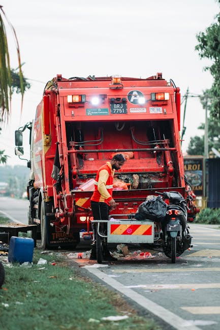 A public waste collection worker wearing a yellow high-visibility vest and black trousers is standing in front of a large red rubbish truck parked on the side of the road. The worker appears to be operating or inspecting the rear loading mechanism of the vehicle, which has a closed compactor area with visible hydraulic components and safety markings. There is a black rubbish bag placed on a metal platform attached to the truck, along with some smaller debris or waste items scattered on the ground nearby. A motorbike is parked close to the work area, with its front wheel partially visible. The truck is situated on a paved urban roadside, with a grassy verge and a few trees in the background. Overhead power lines run parallel to the road, and blurred signage indicates commercial activity beyond the scene, suggesting an environment where private waste collection or on-site clearance may be occurring as part of a rubbish removal service. The overall setting is outdoors during daytime with overcast or diffused lighting, typical of urban waste management operations, with a focus on efficient and safe rubbish collection practices, exemplified by the presence of House Clearance Bow's service setup in the context of alternative waste handling.