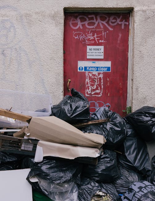 A large collection of overflowing rubbish bins and waste at a designated outdoor waste disposal area in an urban environment. The scene features various materials, including cardboard boxes, paper bags, plastic bags, and discarded packaging, piled on the ground and spilling out of multiple waste containers. One prominent grey bin labeled for mixed paper and cardboard is filled to capacity with loose papers and flattened cartons on top. To its right, a black bin is similarly overflowing with black plastic rubbish bags, some torn open, and debris scattered around. A bright red bin is also present, containing more waste, and next to it are additional boxes and miscellaneous trash, some flattened or crumpled. In the background, a modern building with reflective windows and scaffolding suggests ongoing construction or maintenance work, with bright artificial lighting illuminating the scene. A parked vehicle is visible on the left, and the area is enclosed by metal railings, with a few trees and a paved pavement surrounding the waste collection point. The overall scene depicts an example of unmanaged rubbish accumulation, highlighting the importance of effective waste management and collection services, such as those offered by House Clearance Bow, to prevent illegal dumping and maintain cleanliness in urban spaces.